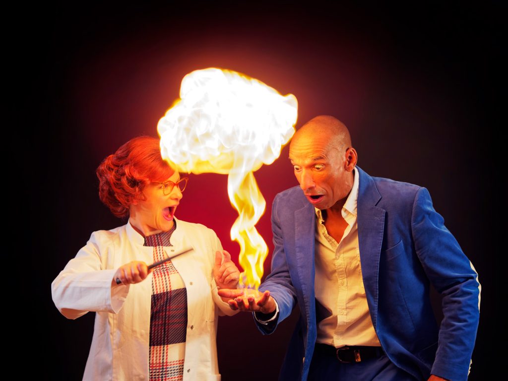 Ein Mann und eine Frau des Ensembles "Die Physikanten" machen erstaunte Gesichter. Die Frau im weißen Kittel erzeugt aus der offenen Hand des Mannes eine Stichflamme .