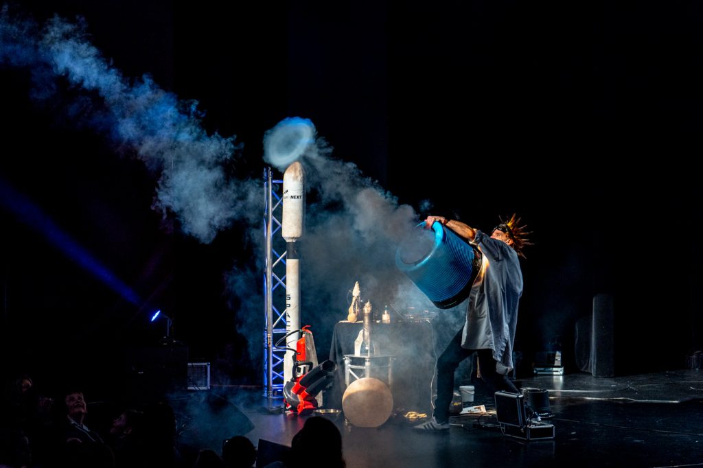 Auf einer Bühne steht ein Mann (der Künstler Professor Bummbastic) und erzeugt mit einem großen zylinderförmigen Objekt, das er in beiden Händen hält, einen Rauchkringel und eine Rauchwolke. Das Bild stammt aus der Wissenschaftsreihe Kammgarn + SWK x Science