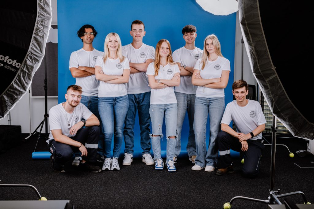 8 Auszubildende der SWK stehen vo einem neutralen Fotohintergrund und tragen alle das weiße Shirt. Das Bild zeigt das Fotoshootinmg der SWK-Ausbildungskampagne 2024
