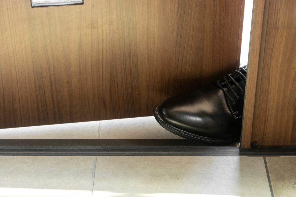 Ein Fuß mit schwarrzem Leder-Herrenschuh wird in eine Tür gestellt, sodass diese nicht geschlossen werden kann (Symbol für aufdringlichen Vertreter)