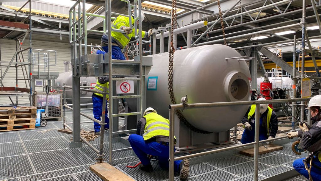 Eine Nahaufnahme einer Kesseltrommel bei der Montage. Einige Arbeiter knien auf dem Boden, einer auf einer Arbeitsb&uuml;hne.