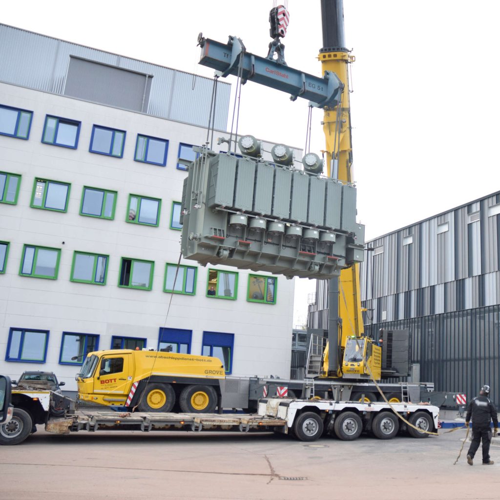 Der Trafo h&auml;ngt frei am Kran und wird hochgezogen. Man sieht das Kranfahrzeug hinter dem Tieflader stehen