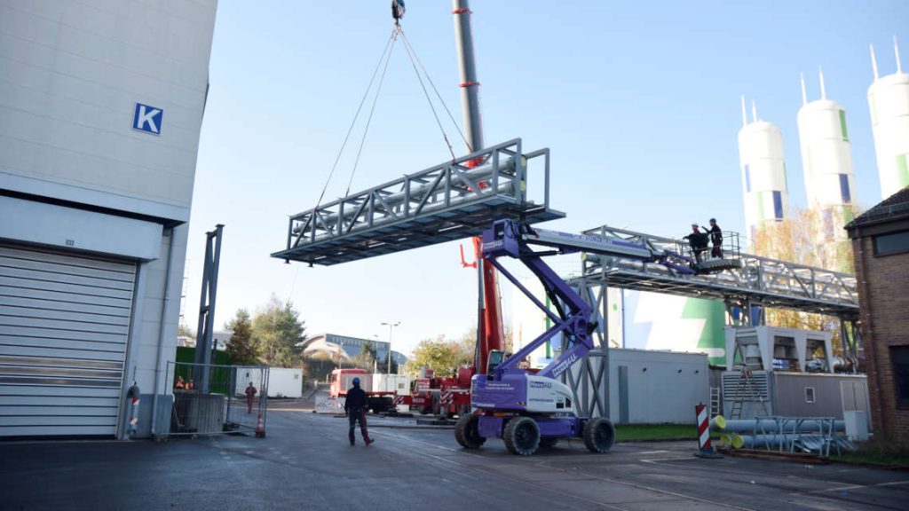 Ein Kran hebt die Rohrbr&uuml;cke in die richtige Position. Arbeiter stehen auf einer Hebeb&uuml;hne und weisen den Kranfahrer ein.