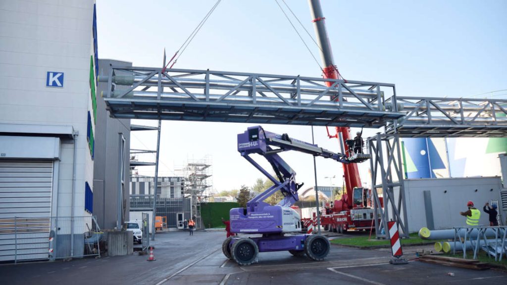 Ein Kran hebt die Rohrbr&uuml;cke in die richtige Position. Arbeiter stehen auf einer Hebeb&uuml;hne und weisen den Kranfahrer ein.