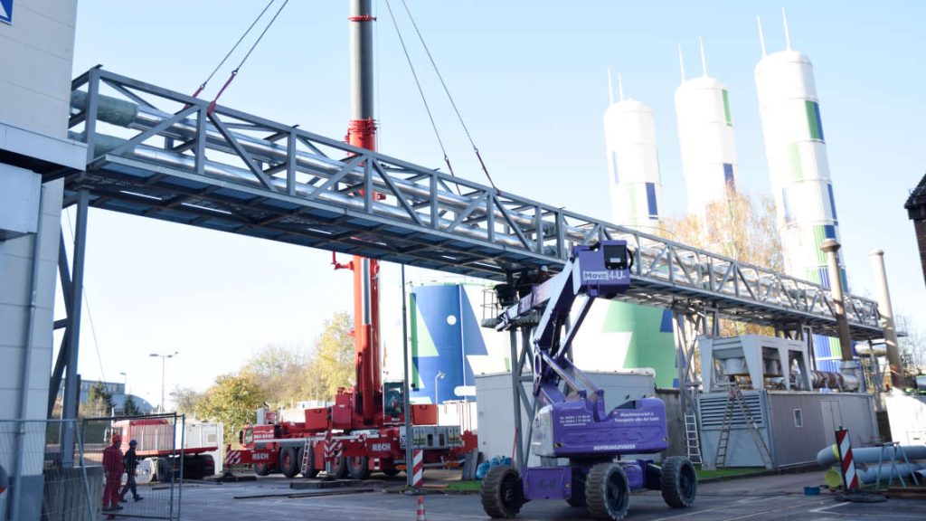 Ein Kran hebt die Rohrbr&uuml;cke in die richtige Position. Arbeiter stehen auf einer Hebeb&uuml;hne und fixieren diese.