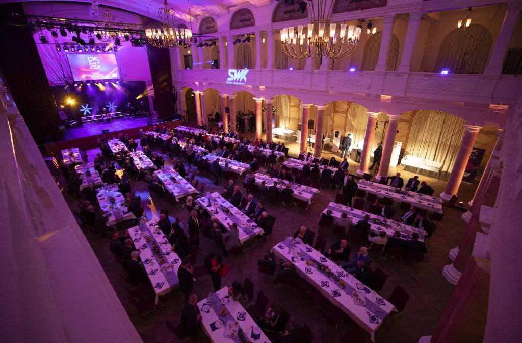 Ein blau und Lila illuminierter Raum in dem 20 Tische stehen an denen Gäste der Veranstaltung sitzen. Auf der Bühne läuft das Programm