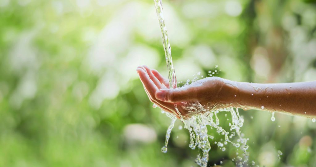 Abgebildet ist eine nach oben geöffnete Hand, die in einen Wasserstrahl gehalten wird.